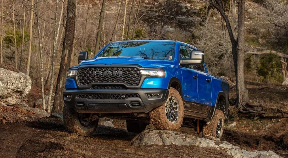 A blue 2025 Ram 1500 Rebel off-roading over muddy rocks.