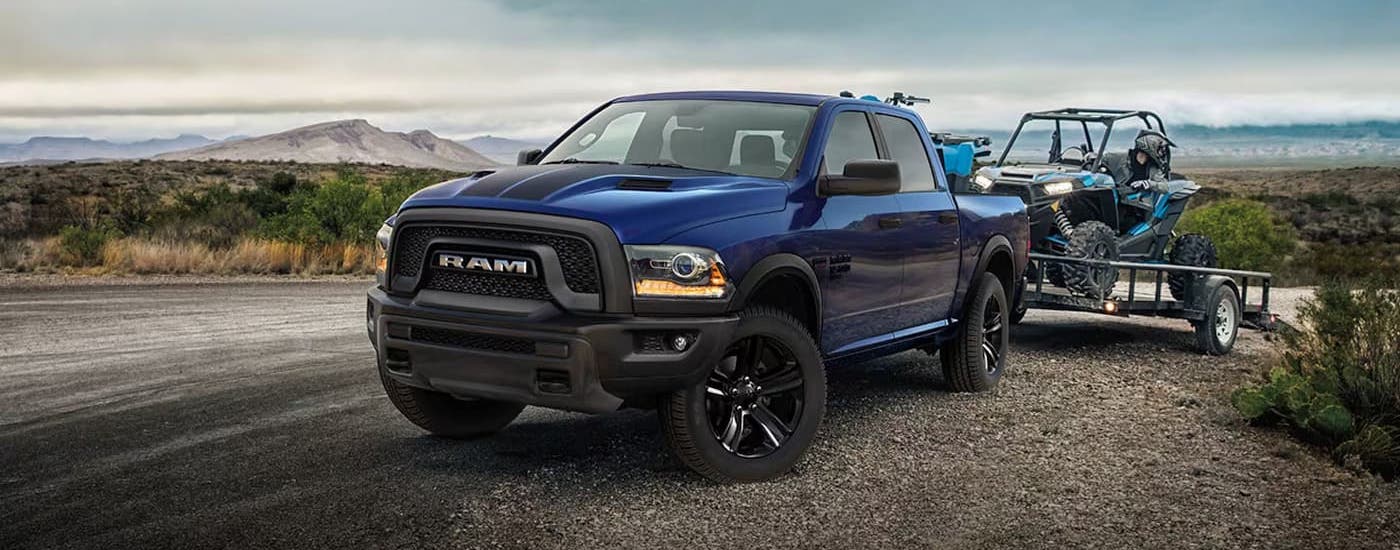 Left angled view of a blue 2024 Ram 1500 towing an ATV off-road.